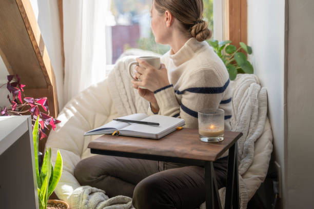 Young woman taking a mental health break to write in her journal stares out the window with coffee in hand stock photo