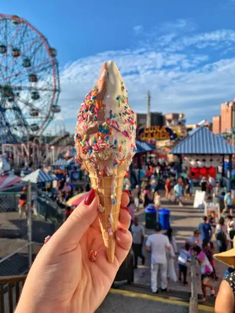 A hand holding an ice cream cone with sprinkles up with Coney Island, New York in the background.