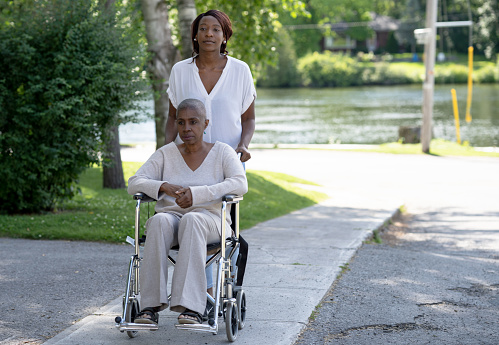 A young woman pushes her Mother, who is undergoing Chemo, in a wheelchair through the neighborhood.  They are both dressed casually and taking advantage of the bright sunny day as they spend time together.
