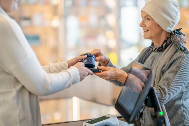 Store Clerk Handing a Customer her Purchase A senior woman with Cancer reaches out for her cannabis product at a legal retailer, as the store clerk passes it to her.  The woman is dressed comfortably in a sweater and is wearing a head scarf as she takes the product from the clerk.. cannabis store photos stock pictures, royalty-free photos & images