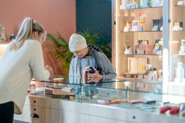 Senior Woman with Cancer Shopping for Cannabis A senior woman with cancer is seen behind the counter of a legal cannabis retailer as she shops for product.  She is gazing down into the glass display cabinet as the store clerk stands on the other side an answers any questions she has.  The woman is dressed comfortably in a sweater and head scarf as she chooses her products. medical cannabis stock pictures, royalty-free photos & images