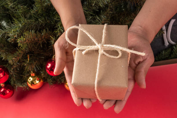 hands with a gift box wrapped in paper with a cord bow, sharing object, studio christmas celebration with tree in the background decorated stock photo