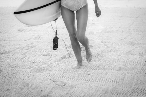 una chica surfera caminando por la playa sosteniendo tabla de surf - desaturado fotografías e imágenes de stock