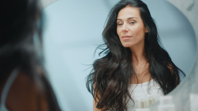 Beautiful Early Middle Aged Woman Looks into Bathroom Mirror Touches Her Lush Black Hair, Admires Her Looks. Concept for Happiness, Wellbeing, Natural Beauty, Organic Skin Care Products