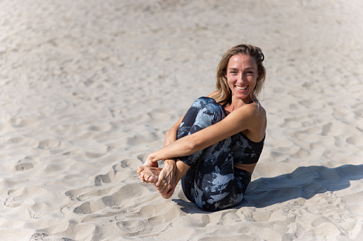 Mid adult woman in yoga outfit sitting on the sand, curled up with knees on chest and holding feet, smiling at camera