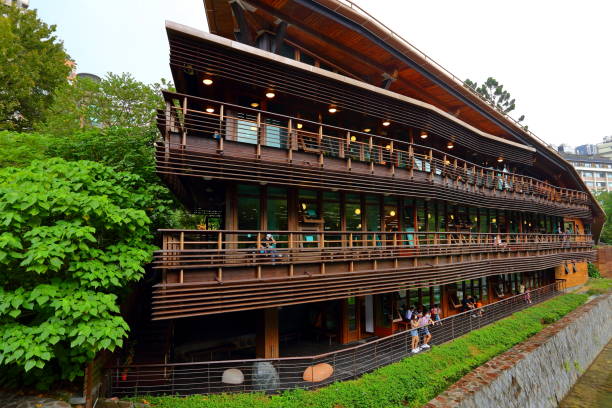 A serene architectural courtyard featuring a wooden boardwalk leading toward a set of escalators and stairs that connect different levels of a red brick building complex. The design blends modern architecture with natural elements, including lush green lawns, tropical trees, and landscaped plantings. A covered walkway on the left provides shade and continuity, while natural sunlight illuminates the open space, creating a sense of openness and tranquility. This type of environment is commonly found in universities, cultural centers, or public institutions, where functionality meets aesthetic appeal. Ideal for themes such as architecture, urban design, campus life, sustainable spaces, or minimalist landscapes.
