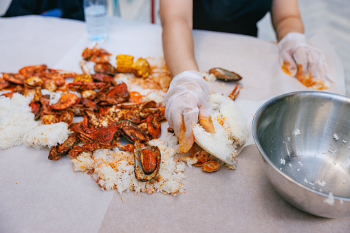 Customer's hands in protective gloves taking rice and seafood served directly on the table, Dampa with mussels, shrimps, squid rings, crab and sweetcorn