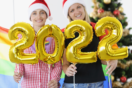 Two women in santa claus hats hold the numbers 2022 against background of New Year tree. Celebrating Christmas concept