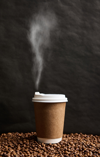 Close-up of a takeaway cardboard cup of hot drink with steam on a surface with scattered roasted coffee beans on a black wall background with copy space for advertisement