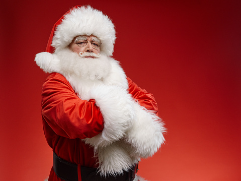 Santa is standing with his arms folded across his chest and looking at the viewer. Santa’s brows are frowned. He is looking displeased. Background is red.
