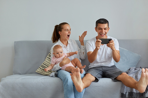 Indoor shot of excited family sitting on sofa in living room, husband holding mobile phone in hands, having excellent news about their winning in lottery, people with infant yelling wow happily.