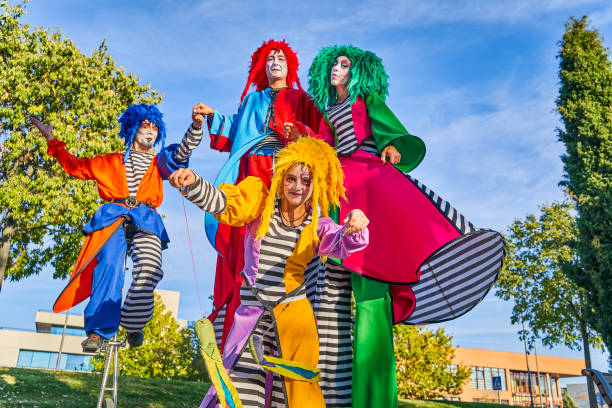 istock Cheerful harlequins on stilts and unicycle in park during masquerade 1353562815
