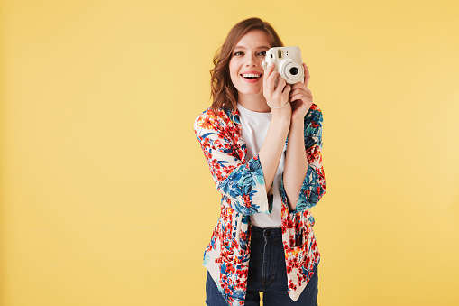 Portrait of young smiling lady in colorful shirt standing with little white camera on over pink background. Pretty girl happily looking in camera