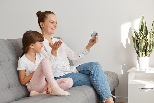 Portrait of beautiful woman with hair bun and her little dark haired daughter with pigtails wearing casual clothing, sitting on sofa in light room and making selfie or having video call.