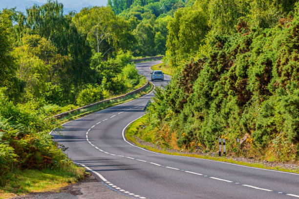 Car on road with curves in Scotland A car drives on a road. A narrow, winding road in Scotland along Loch Ness. Trees and bushes next to the road in summer in sunshine. Traffic signs and guard rails driving winding road stock pictures, royalty-free photos & images