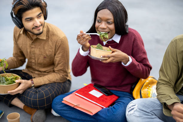 Multiracial students sitting and eating salad during lunch on asphalt at university campus. Concept of education and learning. Idea of student lifestyle. Young smiling friends at food break