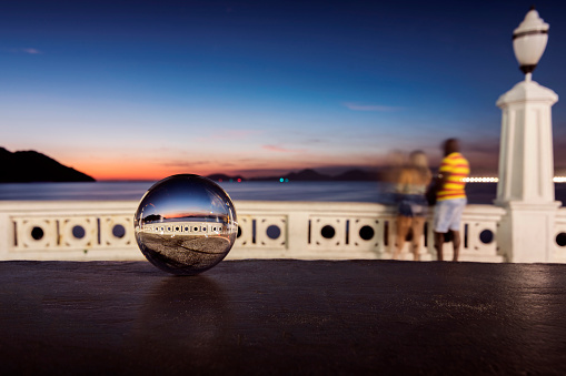 City of Santos. Glass ball in focus reflecting a couple near the wall watching the sunset on the edge of the city. Background with blurred landscape.