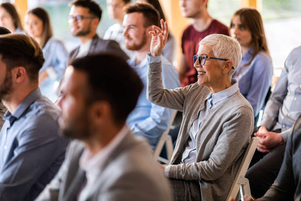 May I ask something? Happy senior businesswoman attending a seminar with her colleagues while raising her hand to ask a question. attending photos stock pictures, royalty-free photos & images