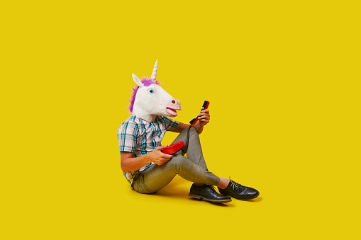 a man wearing a unicorn mask is talking on the phone, using a colorful red landline telephone, sitting on a yellow background with some blank space around him