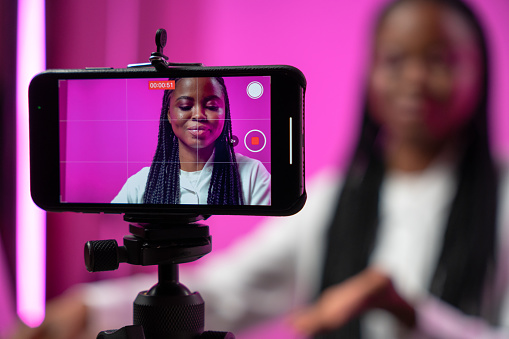A backstage from the filming of a video blog in a home studio with a pink background and a smart phone screen with a dark-skinned blogger on it in the foreground of a picture