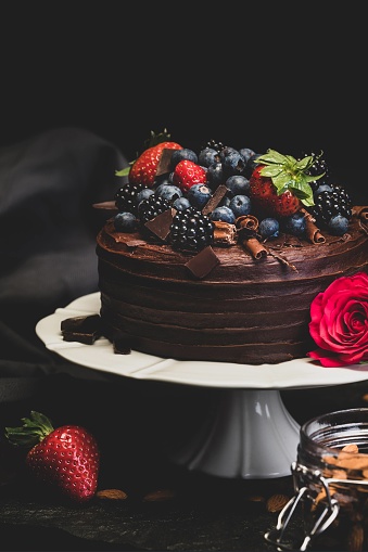 Delicious Double Chocolate Cake Dessert topped with Chocolate Flakes and Summer Fruits, including blackberries, strawberries and blueberries. Styled on a white cake stand with a rose and jar of almonds against a dark background on stone