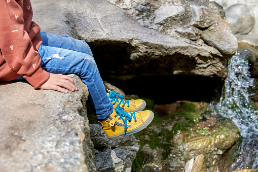 legs of a little boy sitting on a ravine of waterfall of a creek, wearing jeans and yellow boots. Danger, unsafe . Horizontal