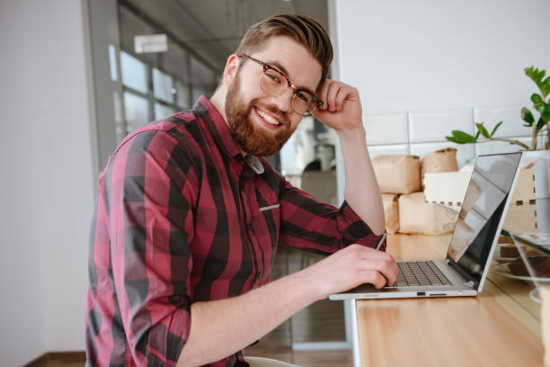 istock Smiling bearded young man in glasses using laptop 1353604111