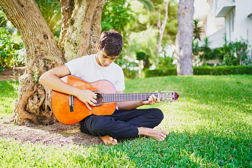Full body of focused barefoot teen male playing music on acoustic guitar in lush verdant backyard