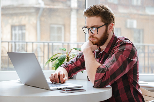 Image of amazing serious bearded young man wearing glasses sitting in cafe while using laptop computer.