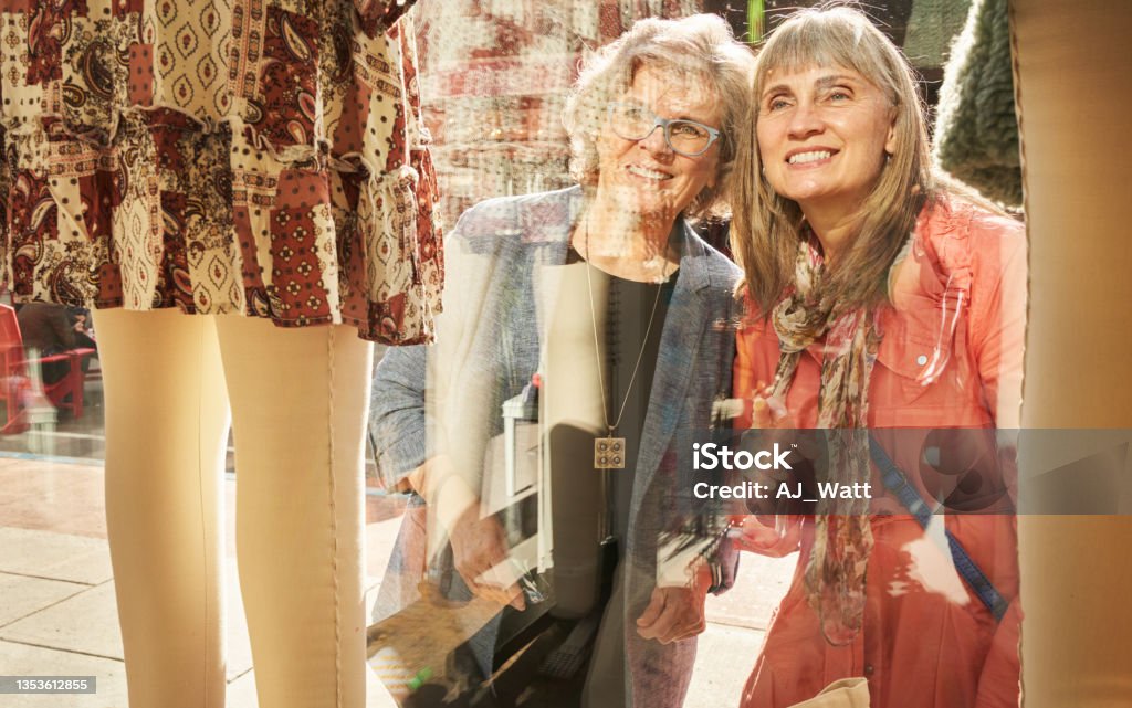 Two mature female friends smiling while window shopping together Two smiling mature female friends looking at a dress while window shopping for clothing together on a asunny afternoon Clothing Store Stock Photo
