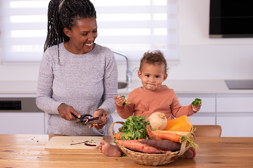 Mother and baby daughter preparing food. Woman peeling a carrot. Baby eating broccoli.