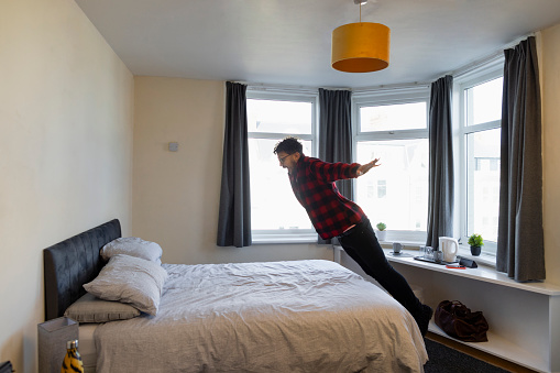 A male hotel guest jumping with his arms outstretched onto a bed in his hotel room in Whitley Bay, North East England after checking in.  He has his mouth open and has an excited look on his face as he jumps.