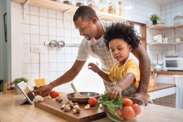 afroamerikanischer kleiner junge, der essen zubereitet, während sein vater auf das digitale rezept schaut und ein touchscreen-tablet in der küche verwendet - garkochen stock-fotos und bilder
