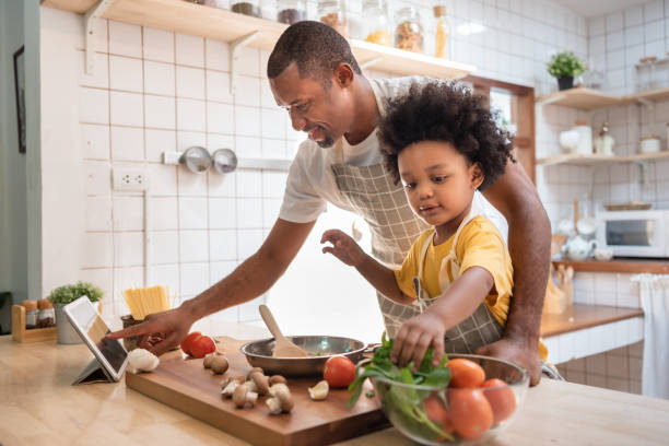 african american little boy preparing food while his father looking on the digital recipe and using touch screen tablet in the kitchen - cozinhar imagens e fotografias de stock