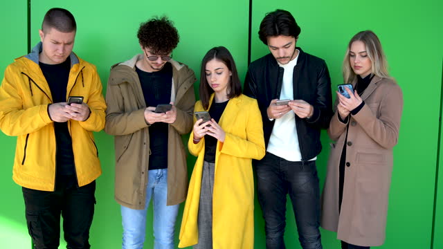 Diverse people guys and girls sitting together holding mobile phones, feel more comfortable chatting online than talking real life. Generation addicted to their smartphones modern technologies concept