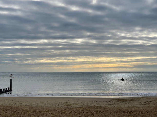 Sea silhouettes of the breakwater groyne with seagull and a lone fisherman in his rowing boat in Poole Bay at sunset on the horizon over water, Dorset, England, UK
