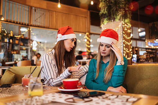 Two best friends with Santa Claus hats sitting in coffee bar or restaurant. One woman is very sad and crying and the other is comforting her. Social or family problems.