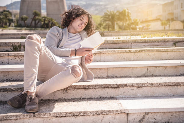 young man sitting on the stairs reads a book outdoor in a city break