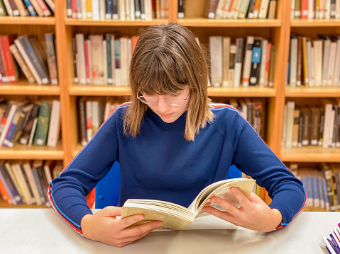 Young teenager sitting on a chair reading a book in the library