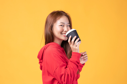 Portrait of a pretty young Asian woman smile and holding takeaway coffee cup wearing red sweater isolated on yellow background. Beautiful young girl enjoys coffee in the morning.