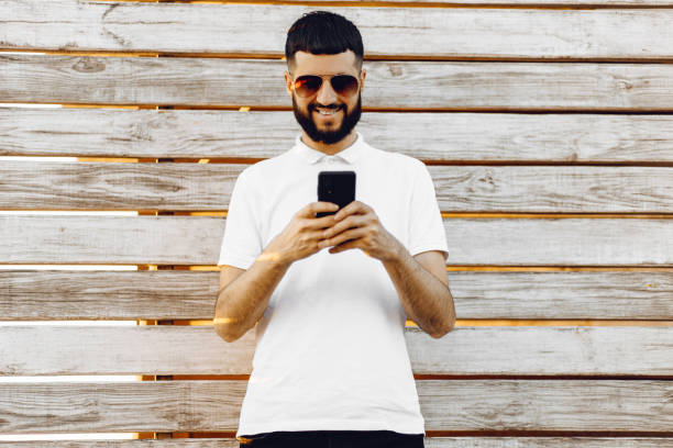 istock Handsome young bearded man using his phone with a smile while standing on a wooden background outdoors 1353656990