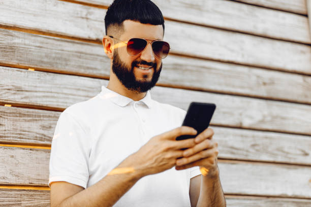 istock Handsome young bearded man using his phone with a smile while standing on a wooden background outdoors 1353657255