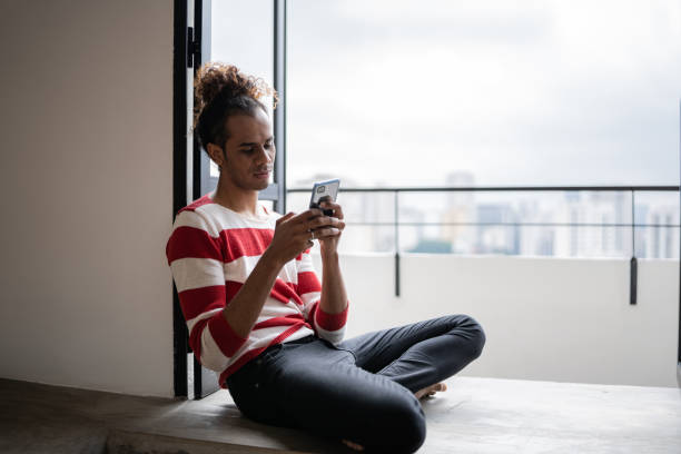 집에서 창 틀에 스마트 폰을 사용하여 지상에 앉아있는 젊은 남자 - telephone window mobile phone addiction 뉴스 사진 이미지