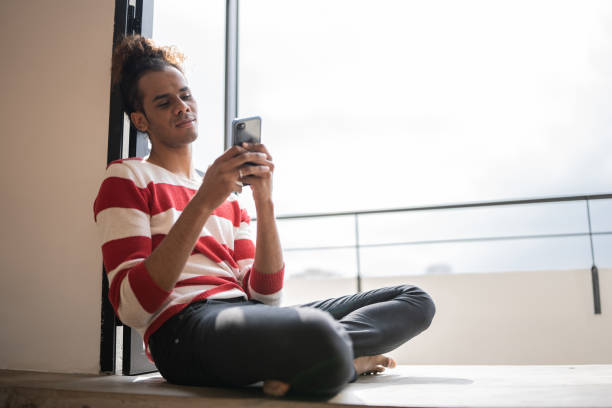 istock Young man sitting on the ground using smartphone on the window sill at home 1353661575