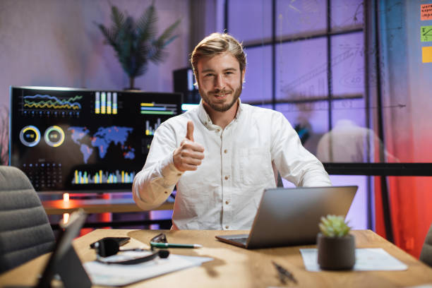 istock Smiling man showing thumb up while sitting at office desk 1353667969