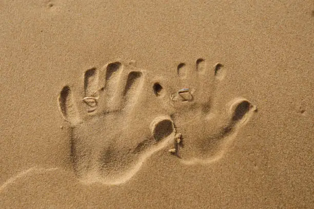 Photo of Two left handprints on the sand with wedding rings on the summer beach. Newlyweds hands shapes