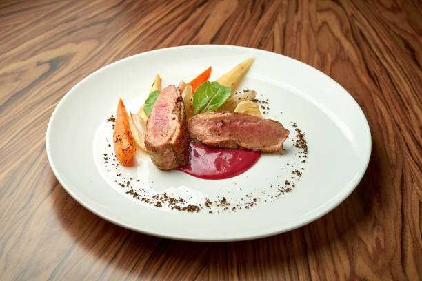 филе утки с запеченными овощами и красным соусом в белой тарелке на деревянном фоне - duck breast стоковые фото и изображения