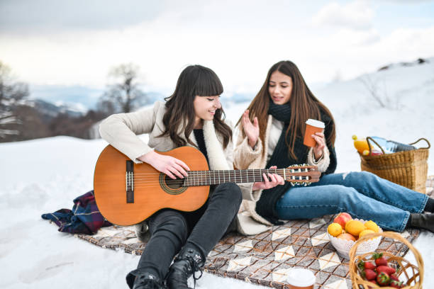 istock Smiling Females Playing Guitar While Drinking Coffee During Snow Picnic 1353673713