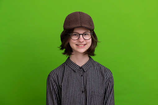 Teenage girl with cap and glasses on green background, looking at camera and smiling.