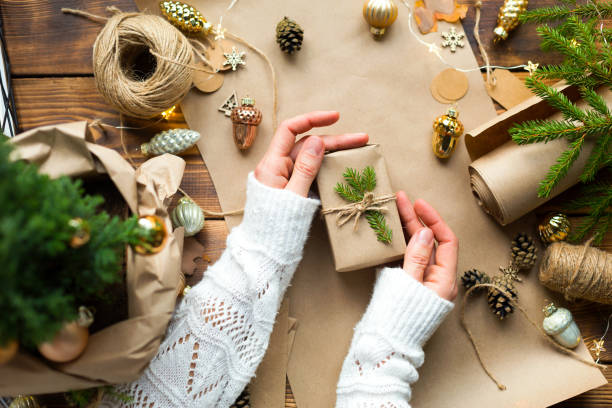 istock Hands in a white sweater pack a gift for Christmas and new year in eco-friendly materials: kraft paper, live fir branches, cones, twine. Tags with mock up, natural decor, hand made, DIY. Festive mood 1353675945
